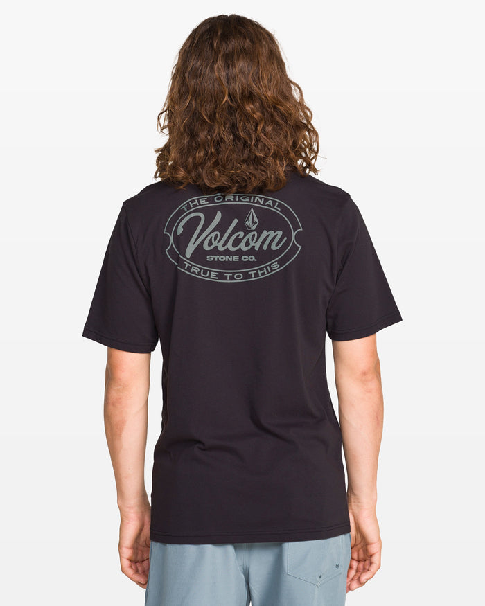 A person with long brown hair, facing away, wears the Volcom Men's Garaged Short Sleeve Tee in washed black heather with a circular “The Original Volcom Stone Co. True To This” back design, paired with light gray shorts.