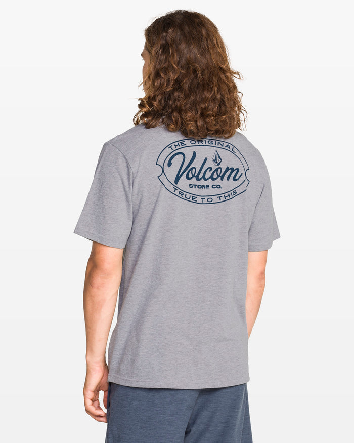 A person with long wavy hair, seen from the back, wears a modern fit heather gray Men's Garaged Short Sleeve Tee by Volcom featuring a large logo and "The Original Volcom Stone Co. True to This" text, paired with blue shorts.