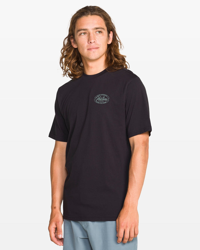 A man with long brown hair models the Volcom Men's Garaged Short Sleeve Tee in washed black heather, featuring a small circular chest logo and modern fit. He pairs it with light blue pants and stands against a plain white background.