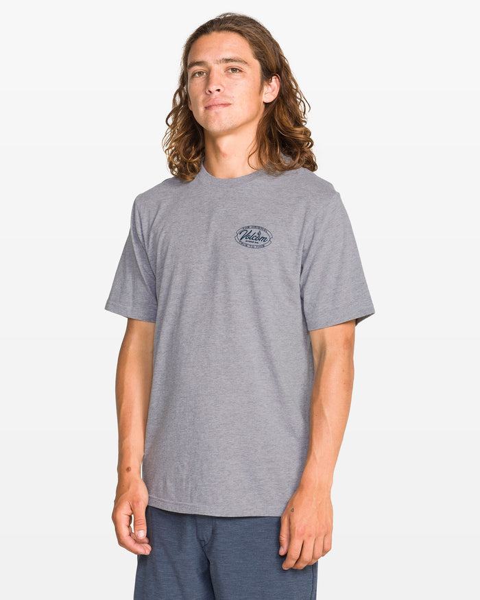A young man with long brown hair stands confidently against a plain background, wearing the Volcom Men's Garaged Short Sleeve Tee in heather gray with a small black chest graphic and blue shorts, smiling slightly at the camera.