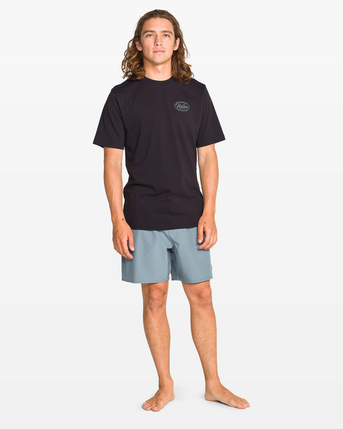 A young man with long brown hair stands barefoot against a white background, wearing the Volcom Men's Garaged Short Sleeve Tee in washed black heather and modern-fit light blue shorts, featuring a small logo on the chest.