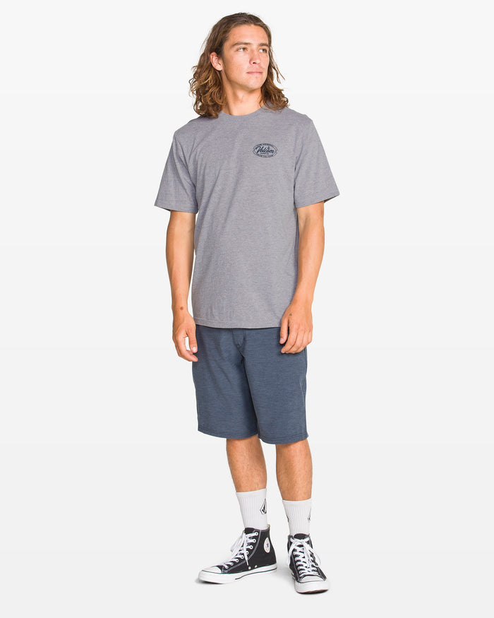A young man with long hair wears the Volcom Men's Garaged Short Sleeve Tee in heather gray, paired with blue shorts, white socks, and black high-top sneakers. He stands against a plain white background, looking slightly to the side.