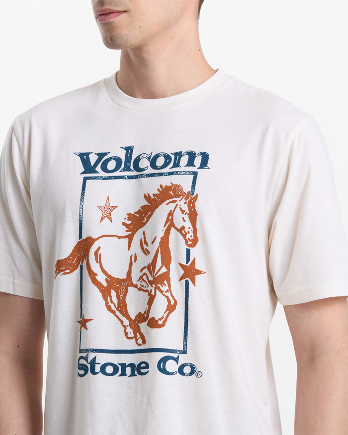 A man wears Volcom's Men's Giddy Up Short Sleeve Tee, a white shirt with a graphic of a running horse, stars, and "Volcom Stone Co." in blue and brown. Only his upper torso and lower face are shown.