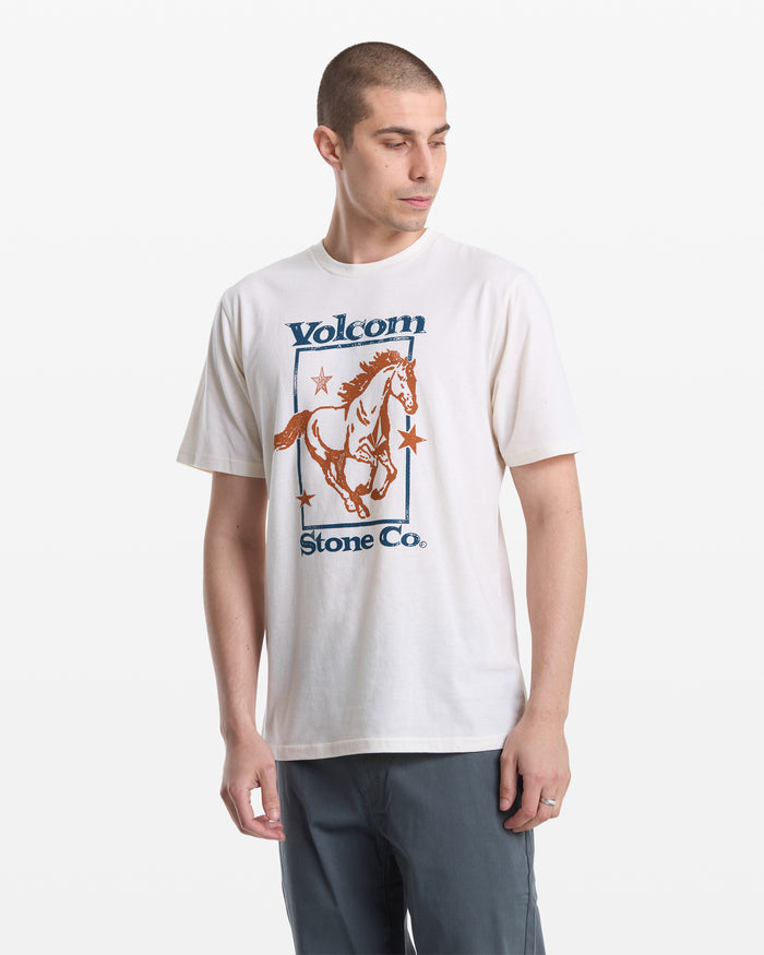 A man with short hair wears the Volcom Men's Giddy Up Short Sleeve Tee, featuring a horse graphic and “Volcom Stone Co.” text in blue and orange, standing against a plain white background.