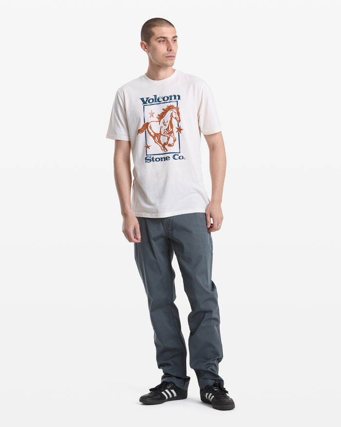 A young man with short hair wears a white Volcom Men's Giddy Up Short Sleeve Tee, gray pants, and black Adidas sneakers. He stands facing slightly to the left against a plain white background.
