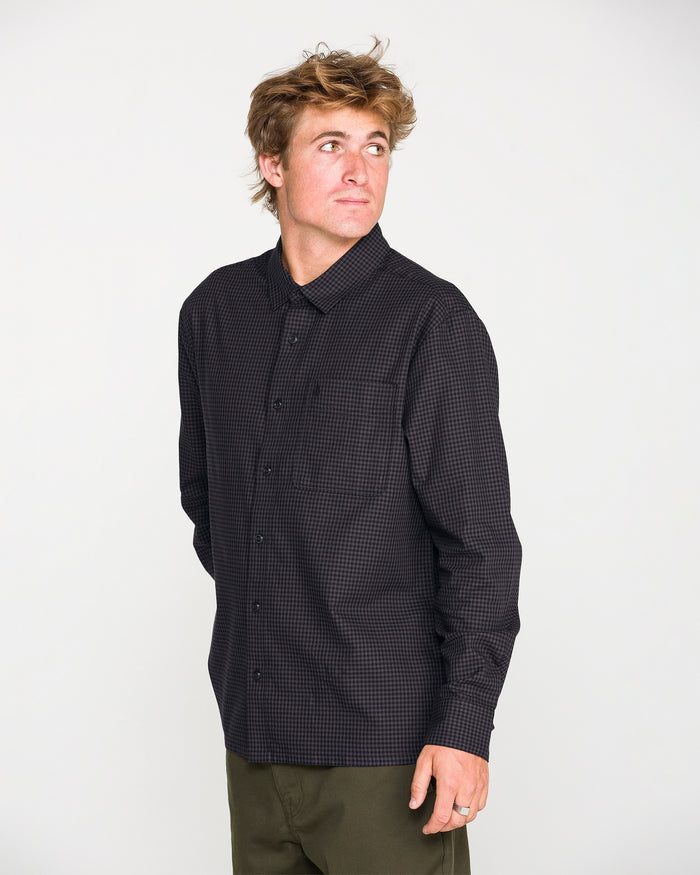 A man with tousled hair wears the Volcom Men's Glasnow Long Sleeve Shirt, a black checkered, yarn-dyed dobby texture button-up, paired with olive green pants as he looks to his left against a plain white background.