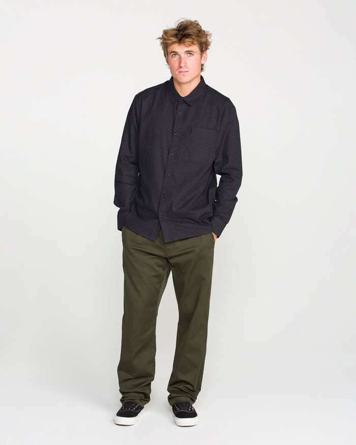 A young man with tousled hair stands against a plain white background, wearing the Volcom Men's Glasnow Long Sleeve Shirt in a dark, boxy fit with a yarn-dyed dobby texture, paired with olive green pants and black sneakers with white soles. His hands are in his pockets as he looks slightly to the side.