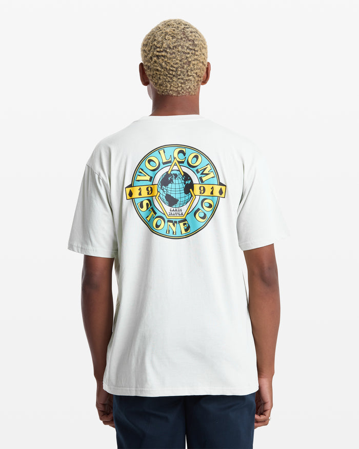 A person with short, light blonde hair is seen from the back wearing the Volcom Men's Globular Short Sleeve Tee, a white shirt featuring a colorful circular logo with a hand holding a globe and "Volcom Stone Co. 1991" text.