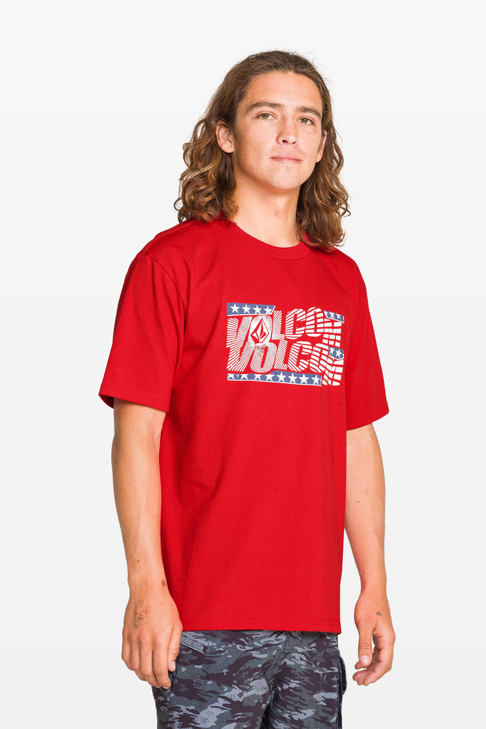 A child with long brown hair wears a Kids' Glory Daze Short Sleeve Tee by Volcom, made from 100% cotton with a patterned logo. They stand against a light background, looking relaxed and wearing dark patterned shorts.