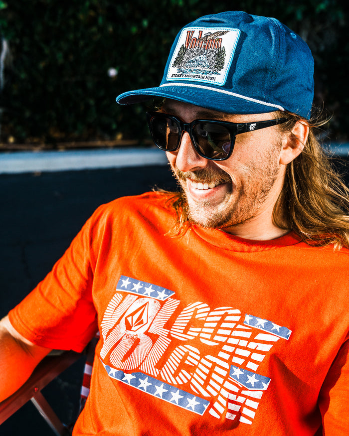 A man with long hair and a mustache, wearing a blue cap, dark sunglasses, and a bright orange Volcom Men's Glory Daze Short Sleeve Tee with stars and stripes, smiles while sitting outside in the sunlight.