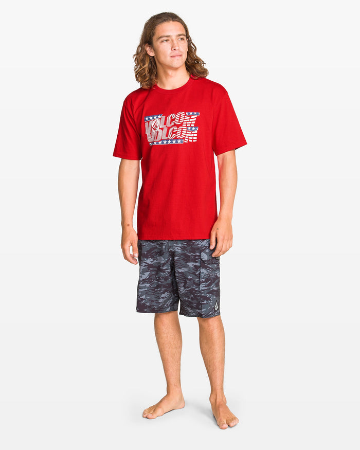 A boy with long hair stands barefoot in a Volcom Kids' Glory Daze Short Sleeve Tee, featuring a classic fit and red 100% cotton fabric, paired with dark patterned shorts against a plain white background.