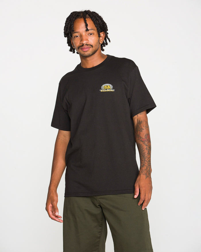 A man with short dreadlocks wearing a black Volcom Men's Golden Bear Short Sleeve Tee made from heavyweight cotton and olive green shorts stands against a plain white background, looking at the camera with a relaxed expression.