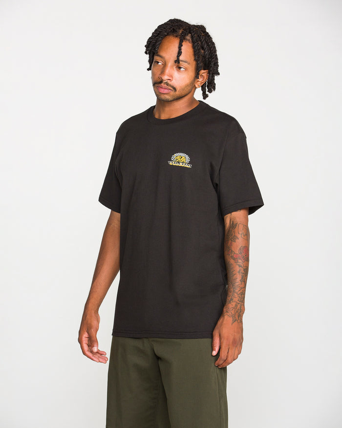 A man wearing the Volcom Men's Golden Bear Short Sleeve Tee—a black heavyweight cotton T-shirt featuring a small round yellow Volcom Stone logo on the left chest—and olive green pants stands against a plain white background.