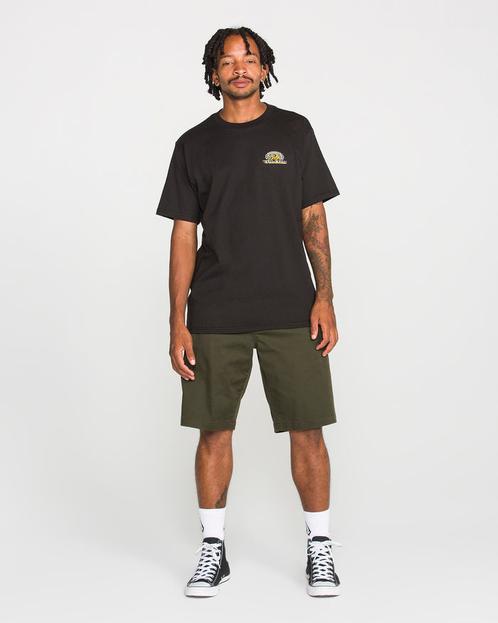 A man stands smiling in a Volcom Men's Golden Bear Short Sleeve Tee crafted from heavyweight cotton, paired with olive green shorts, white socks, and black sneakers against a plain white background.