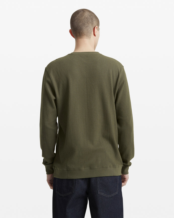 A person with short hair is shown from the back, wearing a Volcom Men's Gridstone Crew Long Sleeve in olive green and dark blue jeans against a plain white background.