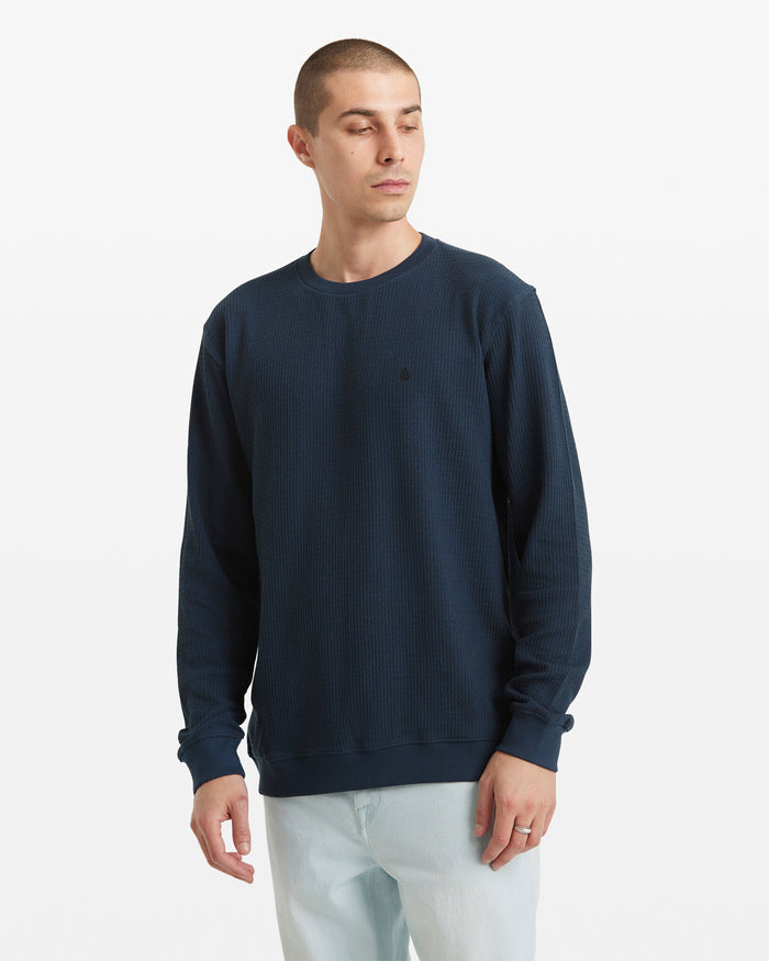 A man with short hair wears a Volcom Men's Gridstone Crew Long Sleeve in navy blue and light blue jeans, standing against a plain white background and looking slightly to his left.