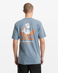 A man wears the Volcom Men's Grindit Short Sleeve Tee, featuring an illustrated welder and "Volcom Workwear Always Grinding Est. 1991" in orange and white on the blue back. Made of 100% cotton, it’s shown from behind against a plain background.