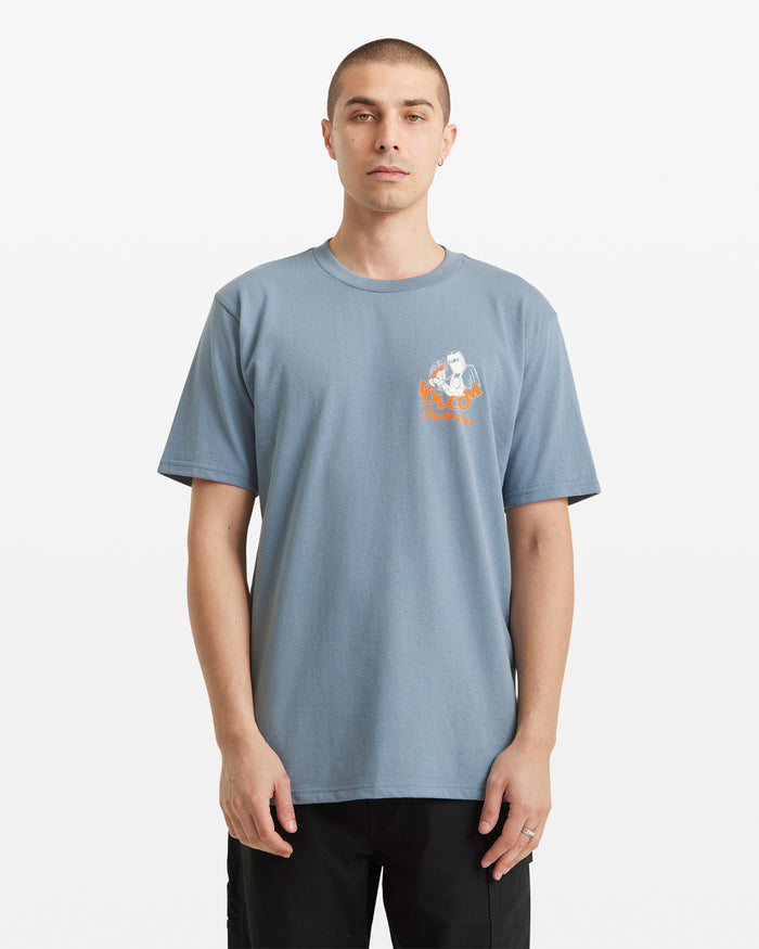 A man wears the Volcom Men's Grindit Short Sleeve Tee, a light blue 100% cotton t-shirt with a small orange and white graphic on the left chest, standing against a plain white background.