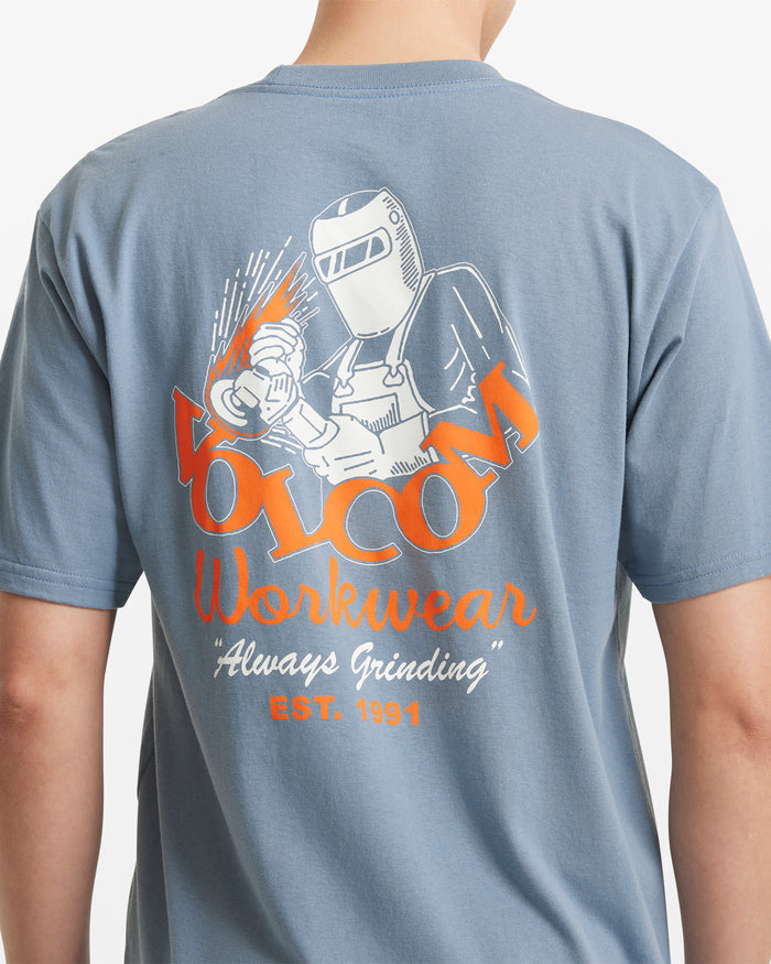 A man wears the Volcom Men's Grindit Short Sleeve Tee, a blue 100% cotton T-shirt with a welder graphic and orange and white "Volcom Workwear, Always Grinding, EST. 1991" text on the back.