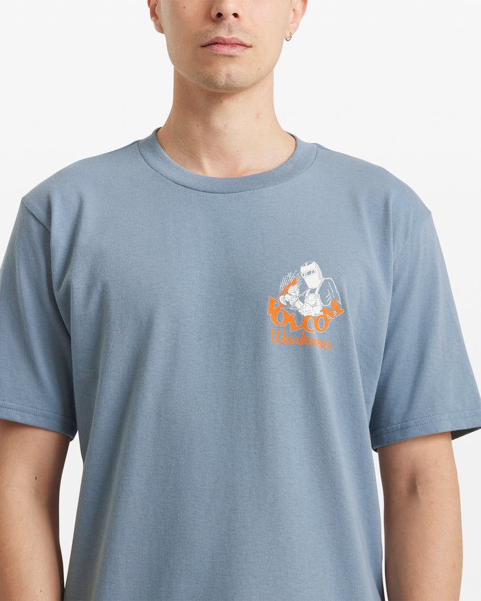 A person wears the Volcom Men's Grindit Short Sleeve Tee, a light blue 100% cotton shirt with a small illustrated worker graphic and “VOLCOM Workwear” in orange and white text on the left chest.