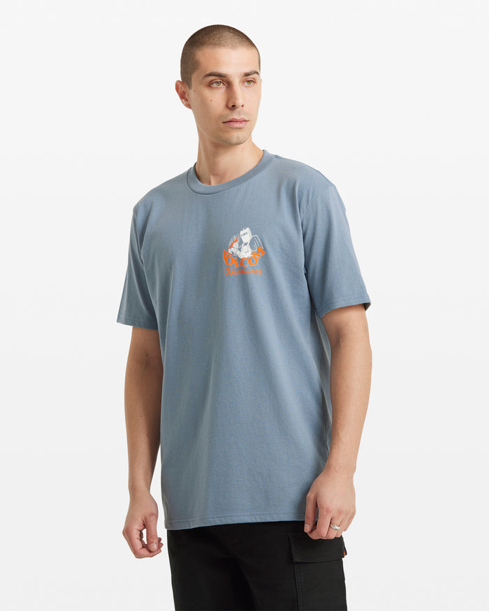 A man with short hair is wearing the Volcom Men's Grindit Short Sleeve Tee, a light blue 100% cotton T-shirt with a small chest graphic and orange text, standing against a plain white background.