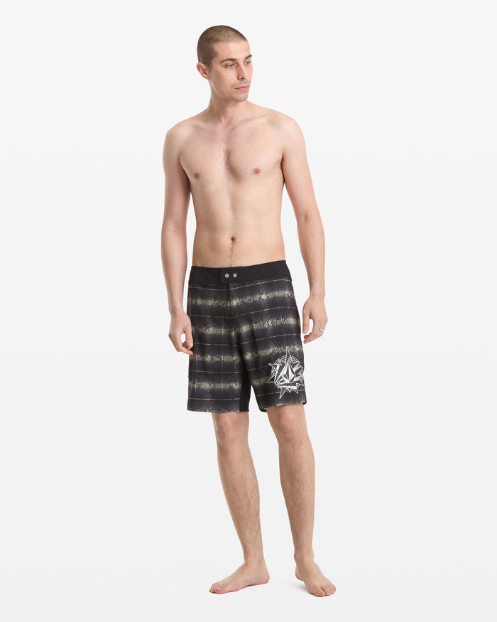 A barefoot man with short hair stands against a white background, wearing Volcom Men's Grunge Snap Mod 18 black and gray board shorts with a white graphic on one leg. He is shirtless and gazing slightly to the side.