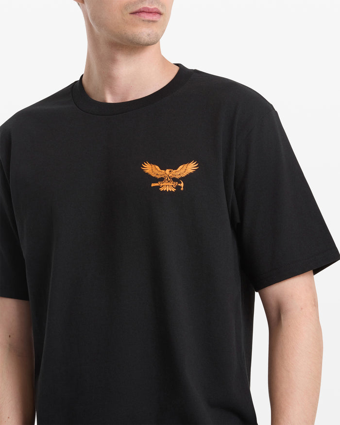 A person models the Volcom Men's Hammered Split Short Sleeve Tee in black, featuring a small gold eagle embroidery on the left chest. The photo is cropped to show the upper torso and part of the face.