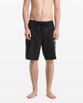 A barefoot man stands against a white backdrop, wearing Volcom Men's Hawaii Top Tier Mod black board shorts with side designs and a drawstring waist. His upper body is unclothed.