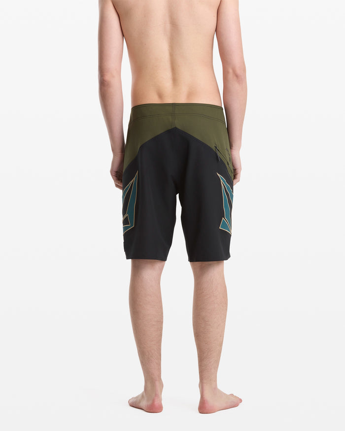 A barefoot man faces away, wearing Volcom Men's Hawaii Top Tier Mod shorts with dark green and black geometric designs, set against a plain white background.