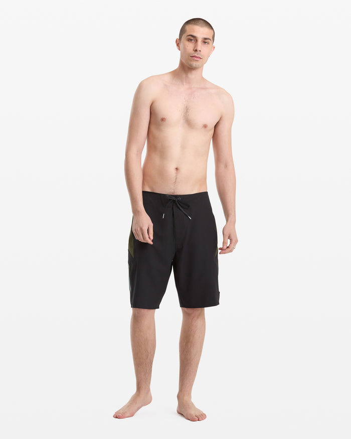 A young man with short hair stands barefoot against a white background, wearing Volcom Men's Hawaii Top Tier Mod black knee-length board shorts with a drawstring and no shirt.