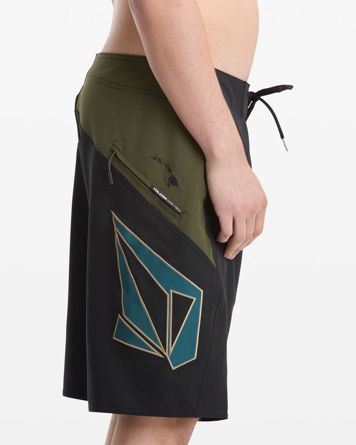 A man wears Volcom's Men's Hawaii Top Tier Mod board shorts in black and olive green, featuring a teal and gold geometric side design, zippered pocket, drawstring waistband; image shows side view waist to knees.
