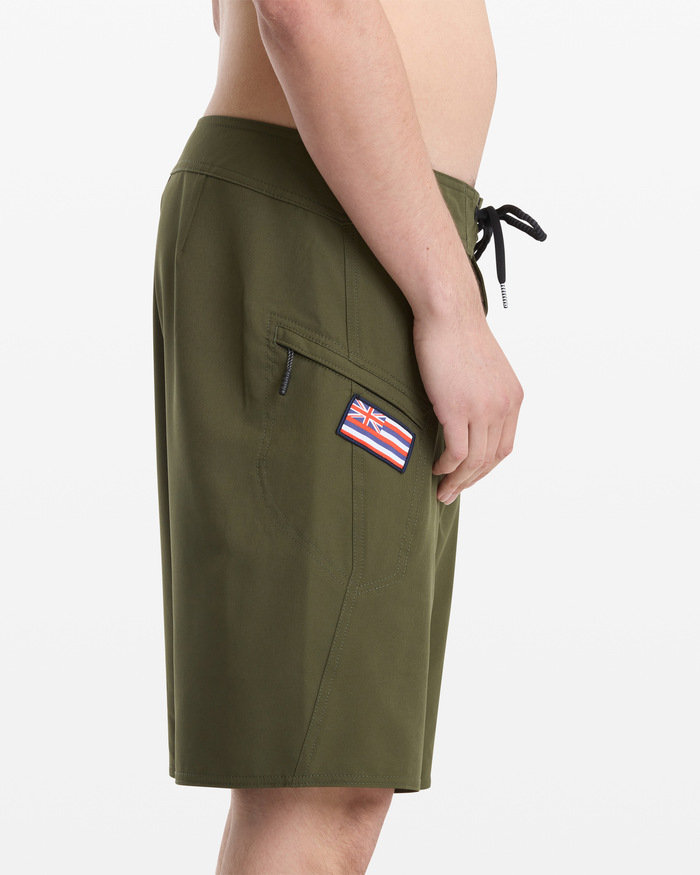 A man is shown from the side against a plain white background wearing Volcom Men's Hawaii Truly Preemo Mod 20 olive green swim trunks with a black drawstring and a Hawaiian flag patch on the side pocket.
