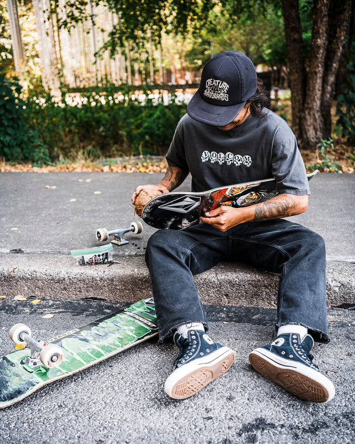 A person in a black hat, Volcom Men's Hedzwillroll Short Sleeve Tee, and black jeans sits on a curb with a skateboard deck. Another board lies nearby as trees and sunlight set the scene for this daily driver moment.