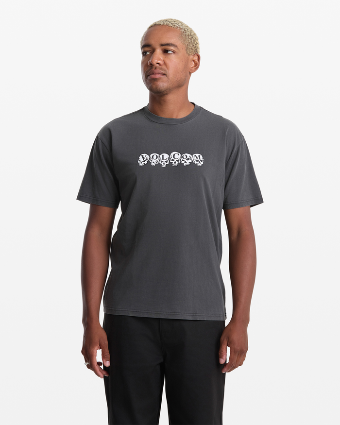 A person with short blond hair models the Volcom Men's Hedzwillroll Short Sleeve Shirt, a dark gray 100% cotton tee with "VOLCOM" in a white skull-style font across the chest, set against a plain white background.