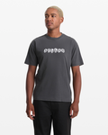 A person with short blond hair models the Volcom Men's Hedzwillroll Short Sleeve Shirt, a dark gray 100% cotton tee with "VOLCOM" in a white skull-style font across the chest, set against a plain white background.