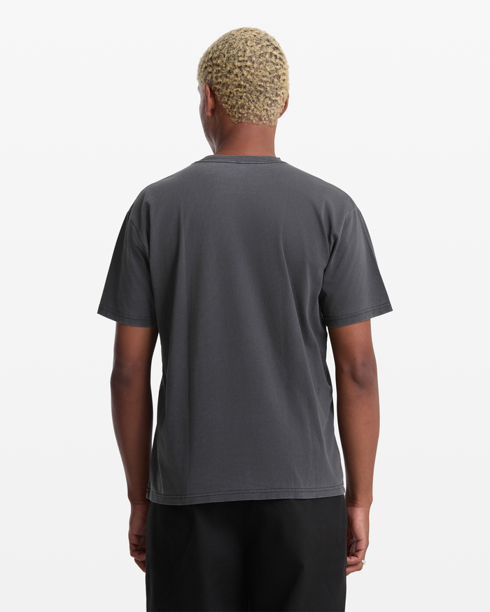 A person with short, curly blond hair stands facing away from the camera, wearing a Volcom Men's Hedzwillroll Short Sleeve Shirt and black pants against a white background.