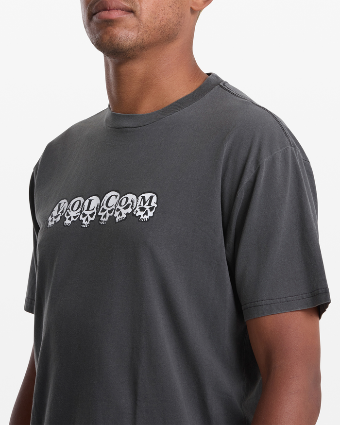 Someone wears the Volcom Men's Hedzwillroll Short Sleeve Shirt, a dark gray 100% cotton tee with "VOLCOM" lettering inside white skull graphics on the chest. The plain white background highlights its perfect daily style.