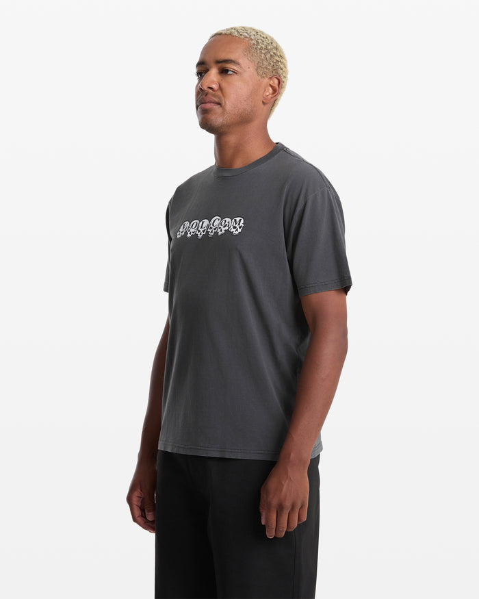 A person with short, bleached blond hair wears the Volcom Men's Hedzwillroll Short Sleeve Shirt in dark gray with a white wavy chest graphic and black pants, standing against a plain white background.
