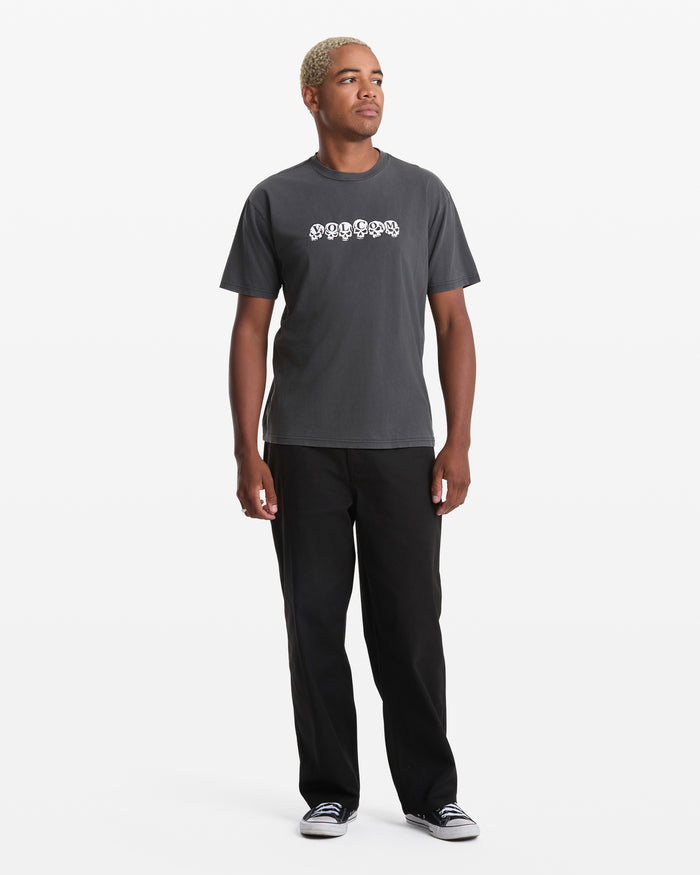 A person with short blond hair wears the Volcom Men's Hedzwillroll Short Sleeve Shirt in dark gray, paired with black pants and black sneakers with white soles, standing against a plain white background.