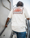 A person with braided hair, walking up metal stairs, is wearing the Volcom Men’s Helios Short Sleeve Tee—a 190g cotton premium shirt featuring a large sun graphic and VOLCOM STORE on the back in soft-hand screenprint.