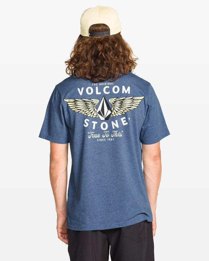 A person with long brown hair wears a backwards beige cap, black shorts, and the Volcom Men's Hi Flyer Short Sleeve Tee in blue, shown from the back against a plain background.