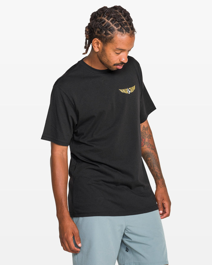 A man with braided hair wears a black Volcom Men's Hi Flyer Short Sleeve Tee made from soft ring spun cotton and light blue shorts, looking down with one arm relaxed at his side against a plain white background.