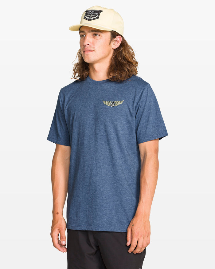 A man with long hair wears the Volcom Men's Hi Flyer Short Sleeve Tee, a modern fit blue ring spun cotton shirt featuring a small winged logo on the chest, styled with black shorts and a light cap against a plain white background.