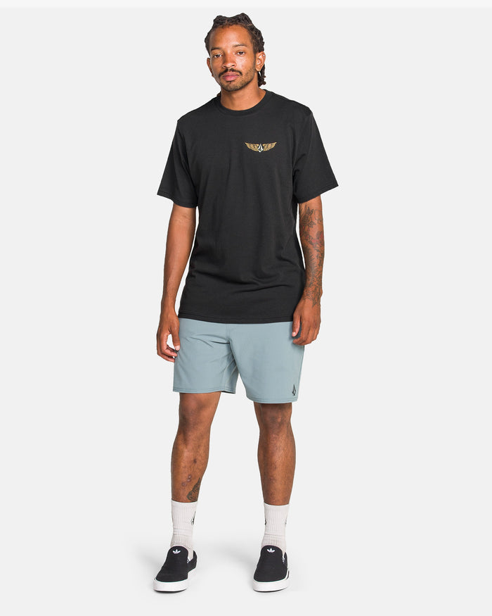 A man with styled hair and forearm tattoos stands against a white background wearing a black Volcom Men's Hi Flyer Short Sleeve Tee, light blue shorts, white socks, and black slip-on shoes.