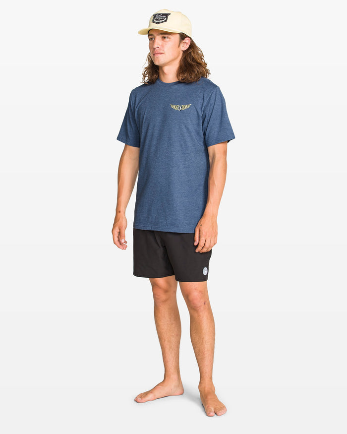 A young man in a beige cap, barefoot, wears the Volcom Men's Hi Flyer Short Sleeve Tee in blue and black shorts while standing against a plain white background.