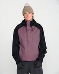 A person is pictured against a plain white background, wearing a beige knit beanie, black gloves, dark pants, and the Volcom Men’s Hossegor 20K Pullover—a purple and black jacket made with a PFAS-free DWR recycled V-Science shell—looking directly at the camera with their hands in their pockets.