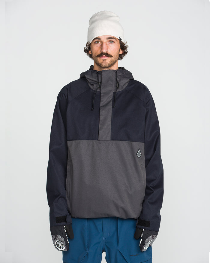A man with a mustache, wearing a white beanie, blue pants, and patterned gloves, stands facing forward against a plain white background. He is dressed in the Volcom Men’s Hossegor 20K Pullover jacket, crafted from recycled V-Science shell in black and gray.