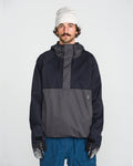 A man with a mustache, wearing a white beanie, blue pants, and patterned gloves, stands facing forward against a plain white background. He is dressed in the Volcom Men’s Hossegor 20K Pullover jacket, crafted from recycled V-Science shell in black and gray.