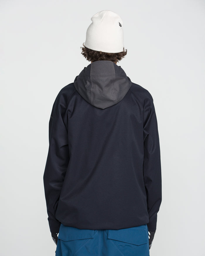 A person wearing a white beanie and blue pants is standing facing away from the camera against a plain light background, dressed in the Volcom Men’s Hossegor 20K Pullover—a dark hooded jacket crafted from a recycled V-Science shell.