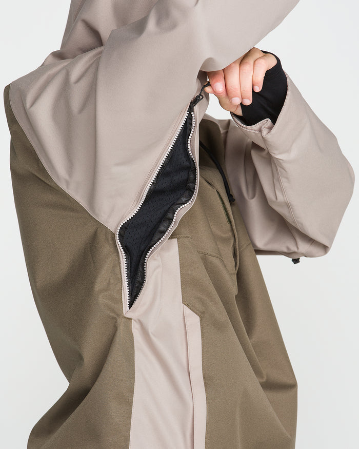 A person pulls up the side zipper of the Volcom Men’s Hossegor 20K Pullover—a two-tone beige and olive jacket with a PFAS-free DWR finish—revealing its black inner lining. Only the torso and part of an arm are visible.
